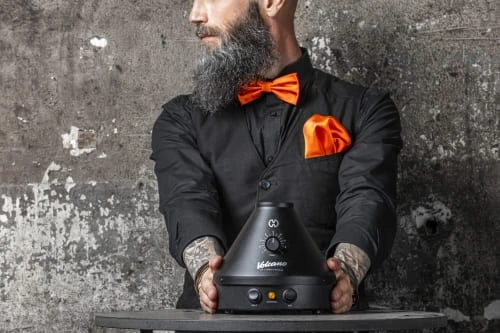 Volcano Classic *ONYX Edition* Ein Mann mit Mattschwarzen Hemd und Orangener Fliege präsentiert vor sich den Volcano Classic Vaporizer in der ONYX Edition